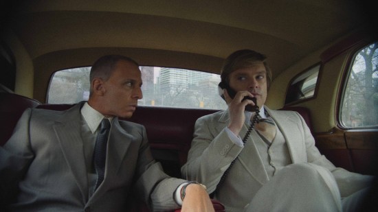 Jeremy Strong and Sebastian Stan sit in character in the back of a limo. Strong glares at Stan, who's on the 1980s car phone.