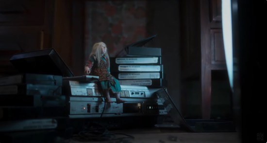 Tiny elderly woman with long hair sits atop a VCR in a dark room, watching TV. Next to her are piles of videotapes with episodes of the fake TV show "Wander to Wonder".