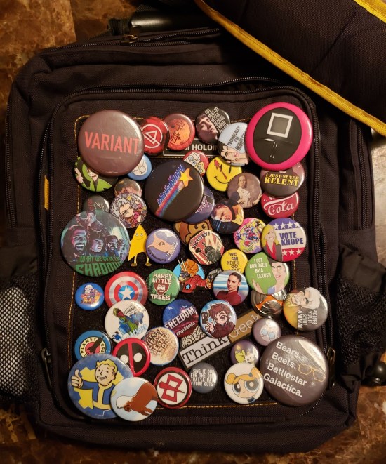 A black satchel covered in dozens of buttons with various pop culture characters, titles and quotes on them.