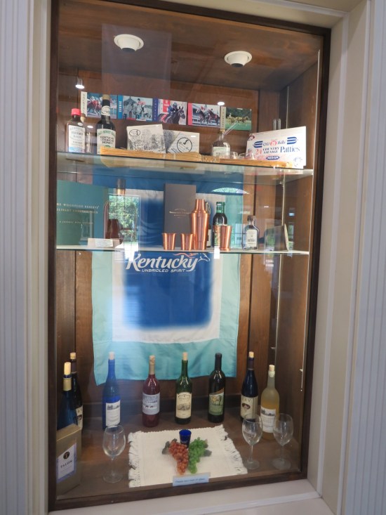 A glass case with lots of bourbon bottles, cups and other relevant implements.