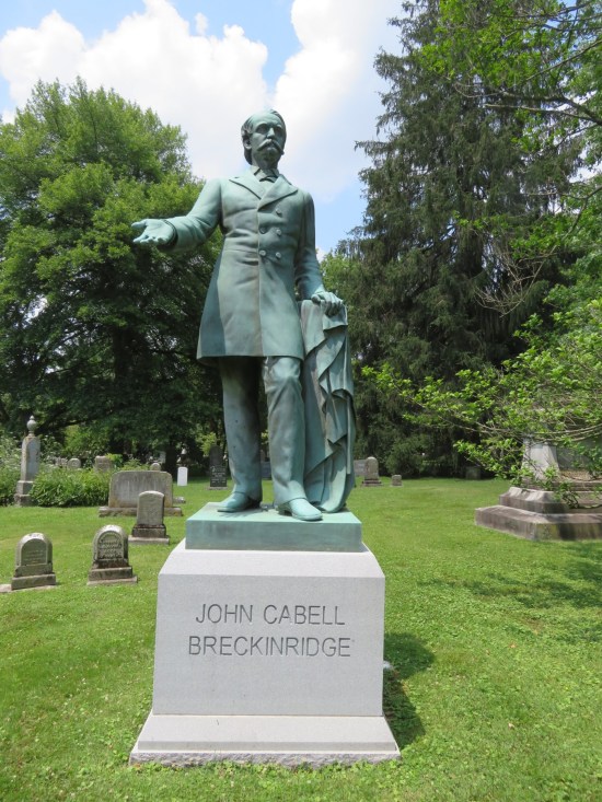 Light blue statue of a white guy in a 19th-century topcoat. One hand is held out palm-up; the other leans on a draped support.