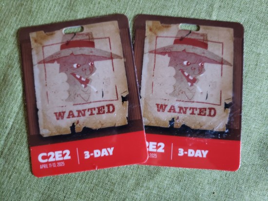 A pair of C2E2 2025 badges on a light green tablecloth. On each is a picture of some thin, deformed animated man with a 1920s Chinatown gangster hat.