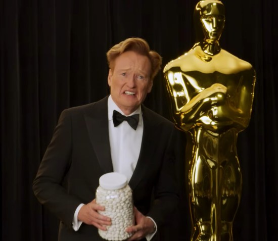 Conan doing photo ops on a black stage with a giant Oscar statue behind him. He's grimacing and holding a large jar of cotton balls or something resembling them.