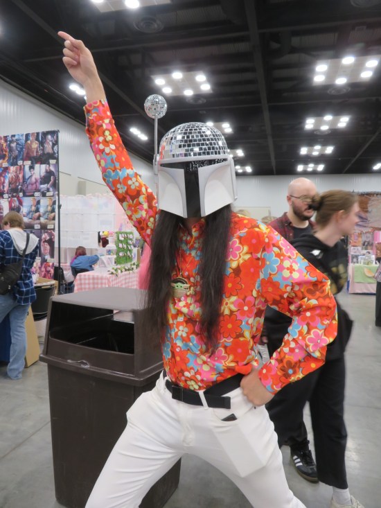 Cosplay: silver Mandalorian helmet with disco ball on top, flowered shirt, white pants, John Travolta pose.