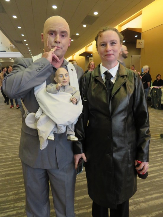 cosplay: Dr. Evil and Frau Farbissina, with Mini-Me doll carried in a sling.