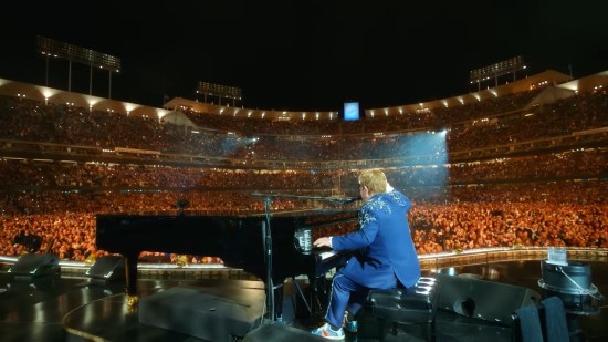 Rear shot of Elton John playing piano onstage at Dodger Stadium at night. Audience is PACKED.
