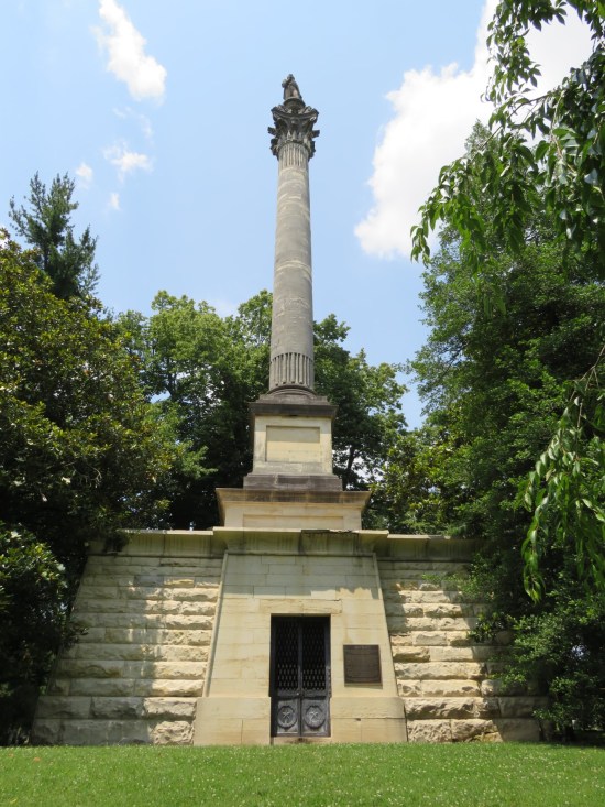 Trapezoidal limestone mausoleum with thin, metal double doors. On the roof is a stone pillar with its own base and a statue on top. Trees surround both sides.