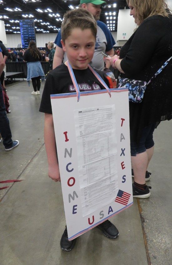 cosplay: young lad wearing sandwich board with two-page Form 1040 and large letters down each side labeling "INCOME TAXES".