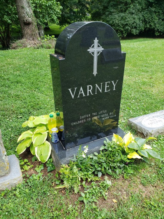 One side of Jim Varney's headstone, with the family name and a cross on it. No decorations are visible on this side except green plants.