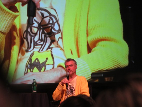 Sean Gunn sitting in front of giant screen also showing him, magnifying his yellow sweater to Godzilla size.