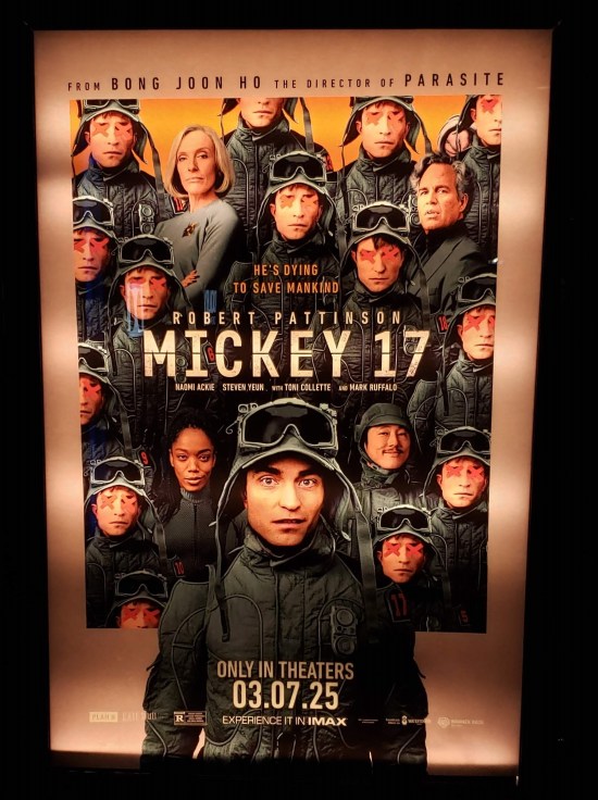 Movie poster for "Mickey 17" hanging in a dark theater with inconsistent backlighting. Poster has multiple Pattinsons surrounding the rest of the cast.