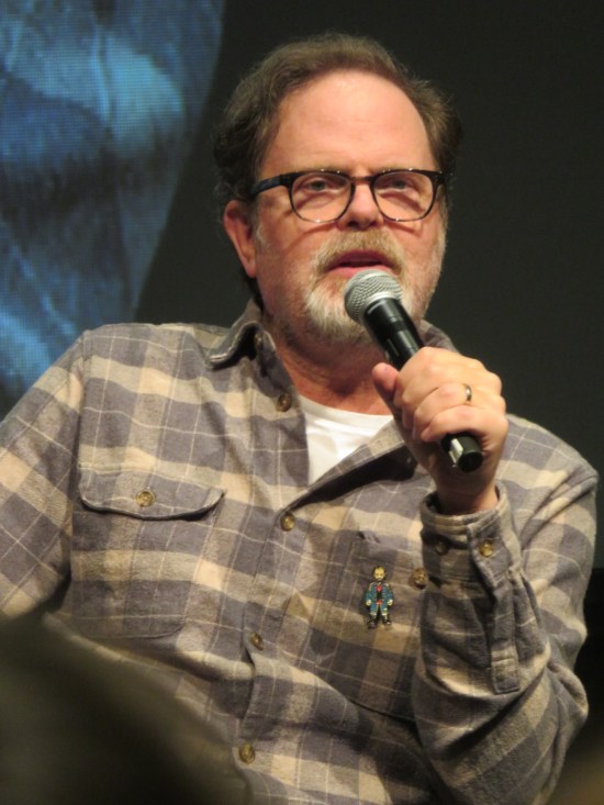Rainn Wilson answers a question on stage in his gray flannel shirt and glasses.