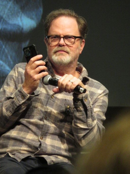 Rainn Wilson holding his phone up to his mic, scowling.