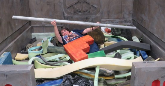 Anne lying under fake space trash, feebly holding up a fake metal pole in ineffective self-defense.