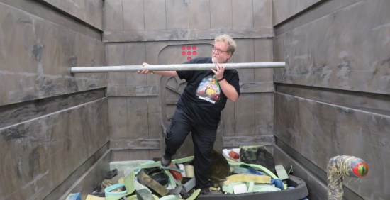 Me propping a fake metal pole against fake metal crushing walls, surrounded by fake garbage.