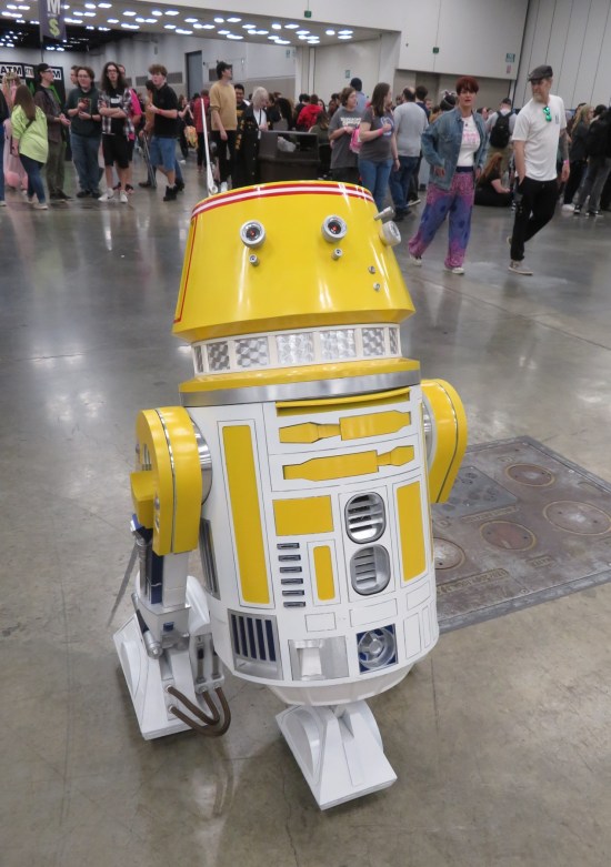 A yellow astromech droid shaped like R5-D4, rolling around the exhibit hall.