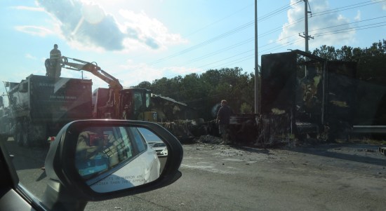 The burnt frame of what used to be a semi trailer. Backhoe and dumptruck are used in cleanup.