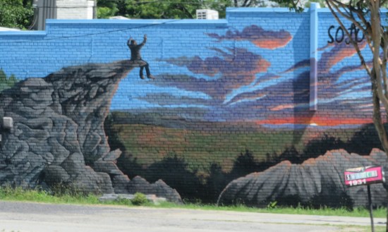 Mural of a guy sitting on a cliff overlooking a green field, watching a pretty sunset with his arms held high.