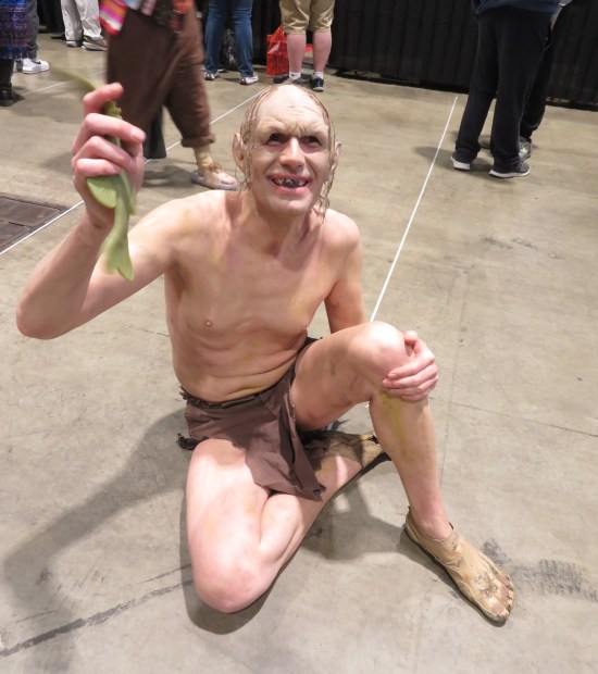 Gollum cosplayer crouched on the convention center's concrete floor, holding a floppy toy fish to you.