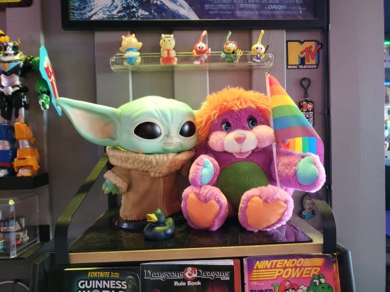 Two stuffed dolls of Grogu and Pancake Popple the same size sitting on a display case. Hanging above them on the wall are five Snorks toys.