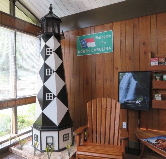 Eight-foot-tall lighthouse model covered in black-and-white diagonal checker pattern. Next to it a wooden patio chair and a TV showing North Carolina tourism stuff.