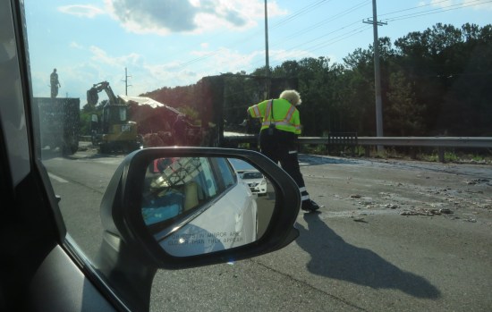 A semi trailer burned to the ground in the right lane. Cleanup crew includes a backhoe and a guy in a fluorescent vest and with a broom.