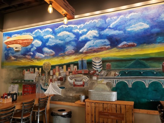 Indoor mural of downtown Knoxville with cloudy skies and a sun on the horizon behind the Great Smoky Mountains.
