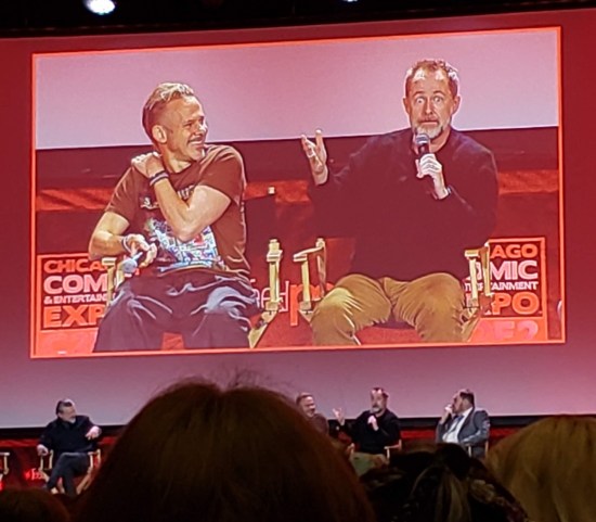 Giant pink-hued ballroom TV screen showing Dominic Monaghan and Billy Boyd seated onstage. Boyd is wide-eyed, Monaghan laughing.