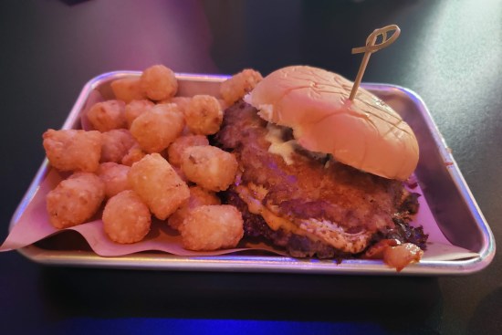 Burger in caption, served in cozy metal tray with tater tots.