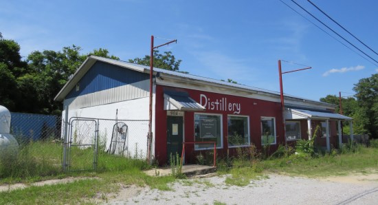 Abandoned red building with "DISTILLERY" sign, lots of unkempt overgrowth in front.