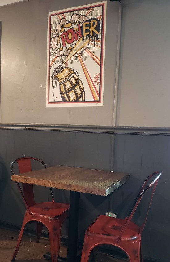 Two red metal chairs at a wooden restaurant table. Hung on the grayish wall behind them is a Pop Art painting of a grenade-shaped perfume bottle spraying a cloud with the sound effect "POWER".