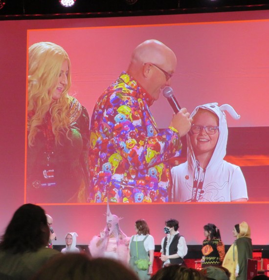 On stage are contestants lined up. On the big screen, our host asks a question of a young girl in a fuzzy bunny suit.