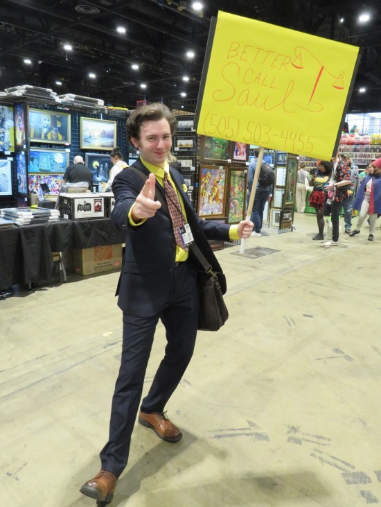 Saul Goodman cosplayer with sign reading "Better Call Saul 505-503-4455" and a hand-drawn set of justice scales.