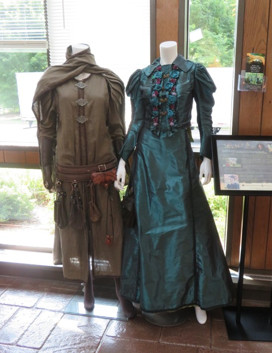 Two outfits hung in a welcome center: one turquoise dress with fancy ruffles and one brown adventurer's outfit with cape and tools dangling from a utility belt.