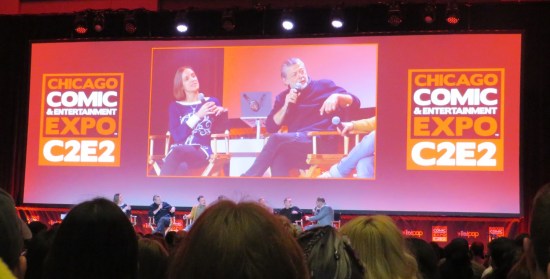 Giant pink-hued ballroom TV screen showing Andy Serkis and a female moderator seated onstage.