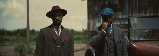 Michael B. Jordan in dual roles as 1932 gangsters, one with a red hat and one with a blue hat.