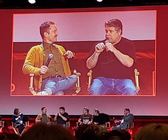 Giant pink-hued ballroom TV screen showing Elijah Wood and Sean Astin seated onstage.