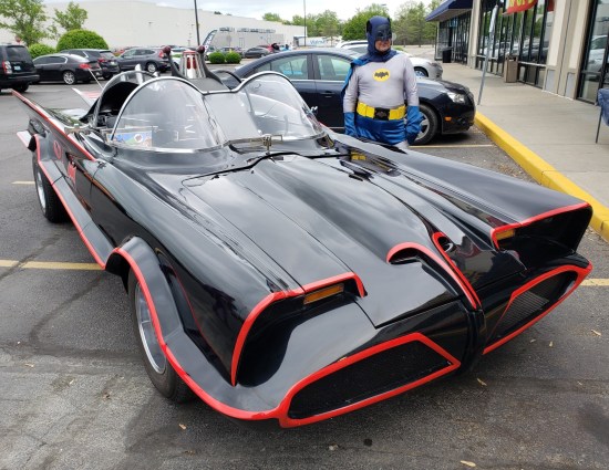 Batman cosplayer standing next to a 1966-style Batmobile, parked outside a strip mall.
