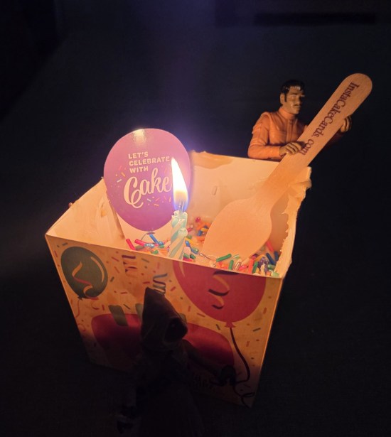 Square cake in a dark room, decorated with lit candle, a cut-out balloon that says "Let's celebrate with cake", and my Willrow Hood action figure pretending to stir it with a wooden InstaCake spoon.