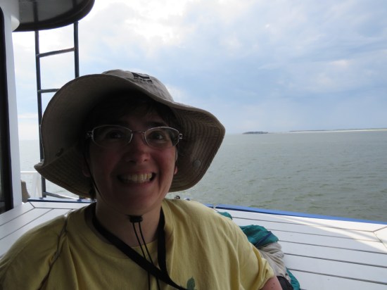 Anne smiling really big on the deck of the ferry. Behind her, the harbor leads out to the Atlantic Ocean. An island is on the horizon.