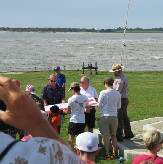 Park ranger holds flagpole rope while guiding civilians stretching an American flag between them.