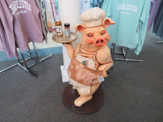 Gift shop statue of a pig standing on two legs, wearing a chef's hat and apron, and holding a serving tray with bottles of barbecue sauce and rub.