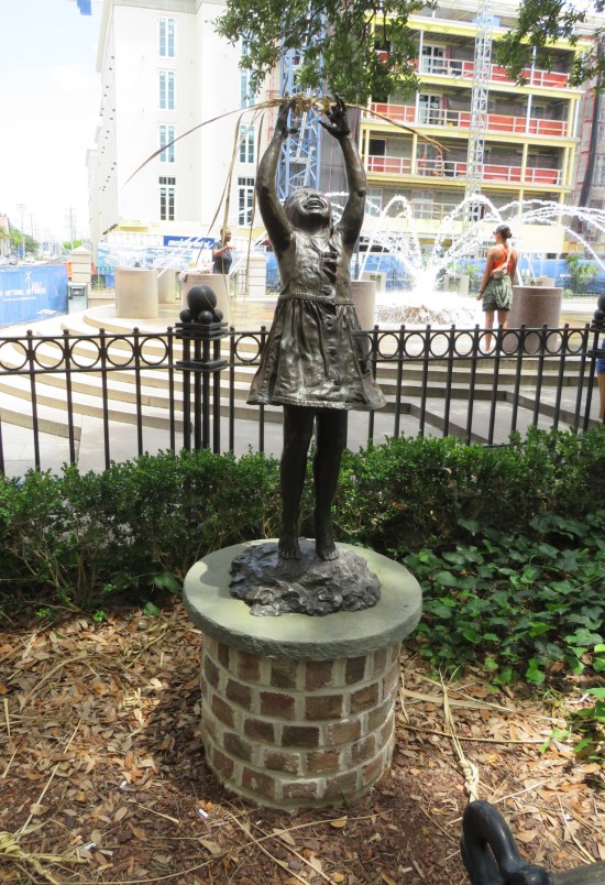 Statue of a Black girl on a pedestal, her arms raised toward the sky as if to grab the stars. Statue is near a public park fence.
