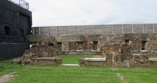 Brick wall with wooden extension on top. Brick benches and shorter walls decay all around in the grass.