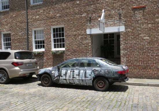 Car covered in graffiti parked on a cobblestone side street.
