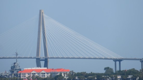 Closer shot of bridge tower with lots of cables emanating from the top. Under the bridge is a white resort with an orange roof and part of a battleship.