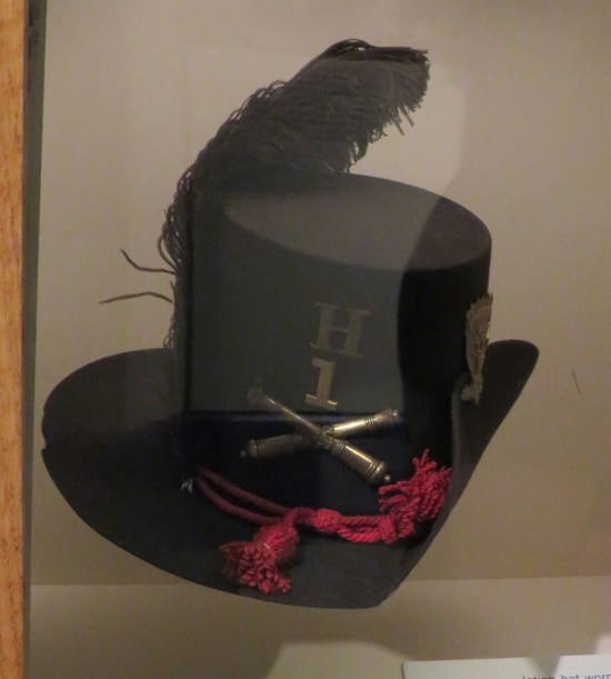 Black top hat with thick red string, bronze pin with crossed firing pins, big black feather, and "H1" on the front.