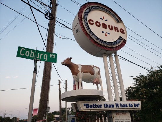 Cow statue mounted on a poled platform next to a big, obsolete Coburg sign.