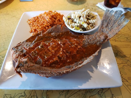 Entire flounder fried to a crisp and spiced. with the fins still attached. Square white plate also has red rice and a tiny bowl of pasta salad.