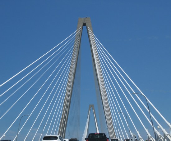 Very tall arch of a white cable-stayed bridge, as seen through our car window with other cars ahead of us. Sky is pretty blue.